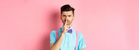 Angry and grumpy man in funny bow-tie shushing at camera, frowning and demand silence, tell to be quiet, scolding someone loud, standing on pink background photo