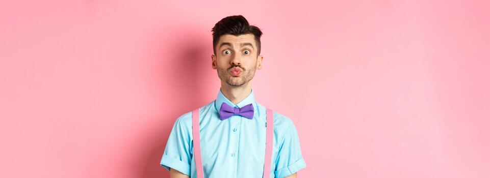 gracioso joven hombre esperando para Beso con fruncido labios y tonto rostro, en pie en romántico rosado antecedentes. concepto de amor y san valentin día foto