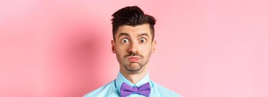 Close-up of confused caucasian guy holding breath, pouting and staring shocked at camera, wearing festive clothes and bow-tie, pink background photo
