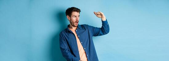 Impressed caucasian guy staring at empty space with big object, shaping large size, standing on blue background photo