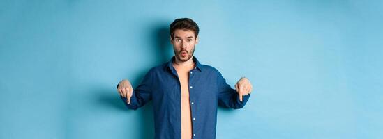 Impressed guy look curious, say wow and pointing fingers down, showing advertisement, standing on blue background photo