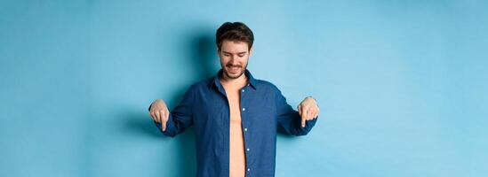 Smiling handsome man with beard, looking and pointing down at banner, checking out special offer, standing on blue background photo