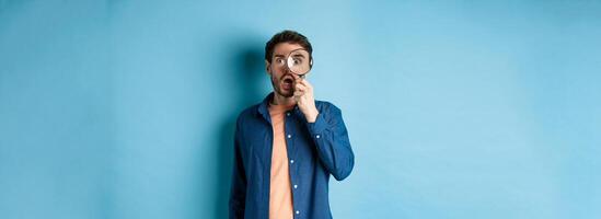 Impressed man look in awe through magnifying glass, drop jaw and stare at camera, standing on blue background photo