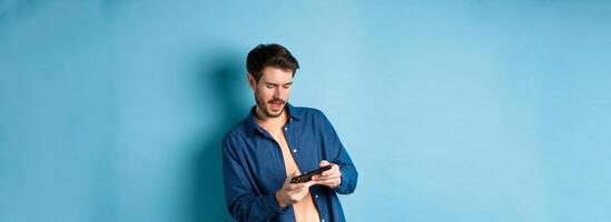 Young guy playing game on mobile phone, tilt body and holding smartphone horizontally, standing on blue background photo