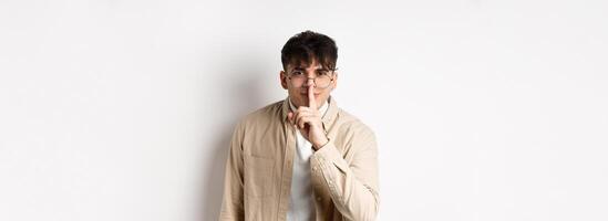 Cheerful handsome man in glasses share a secret, making surprise, lean at camera with hush gesture and gentle smile, telling to be quiet, show taboo sign, white background photo