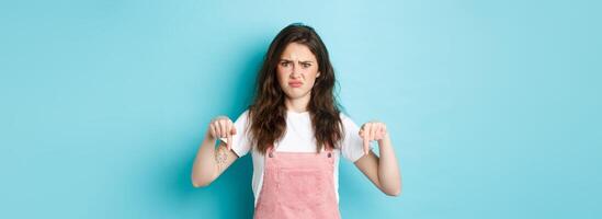 Doubtful young woman frowning and cringe from something bad, pointing fingers down at strange thing, having hesitations, blue background photo