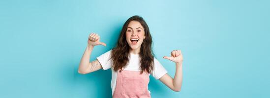 Pick me. Cheerful young girl pointing at herself and smiling, self-promoting, volunteering, looking joyful at camera, standing against blue background photo