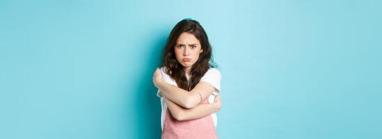 Sad and gloomy young woman pouting, comforting herself with cuddles, hugging her body and looking upset about something unfair, standing against blue background photo