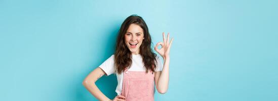 Confident sassy girl showing okay sign and say yes, smiling pleased and determined, approve good deal, praising nice choice, like something good, standing over blue background photo