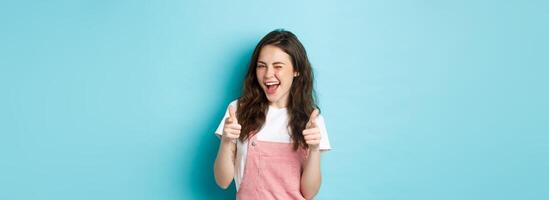 You can do this. Beautiful cheerful brunette girl winking and smiling, pointing fingers at camera, praising good job, inviting to event, standing against blue background photo