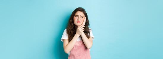 Portrait of frowning girl touching cheek, having toothache, looking at upper left corner stomatology clinic banner, suffering tooth decay, standing against blue background photo