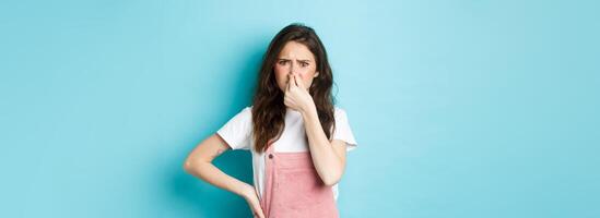 Portrait of bothered cute girl shut nose and frown from bad smell, smelling terrible reek, something stink, standing against blue background photo