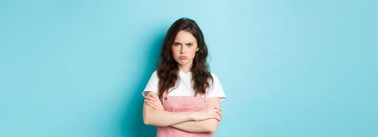 Portrait of angry and upset sulking girl, pouting about unfair situation, frowning and staring at camera, cross arms on chest defensive, standing over blue background photo