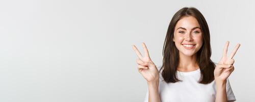 Portrait of beautiful and happy brunette girl smiling and showing peace gestures, standing white background photo