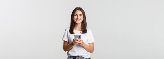 Attractive smiling brunette girl texting message, using mobile phone and looking at camera photo