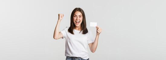 Rejoicing beautiful girl showing credit card and triumphing, fist pump happy, white background photo