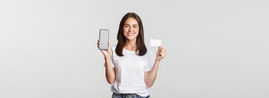 Attractive smiling girl looking satisfied and showing credit card, mobile phone screen photo