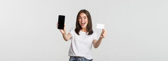 Attractive smiling girl looking satisfied and showing credit card, mobile phone screen photo