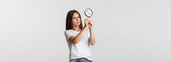 Serious-looking attractive young woman searching for something, looking through magnifying glass, white background photo