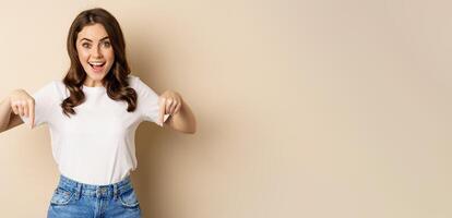 Smiling happy girl pointing fingers down and showing banner, company logo below, click link, standing over beige background photo