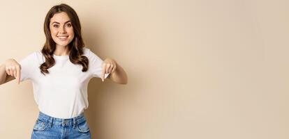Smiling happy girl pointing fingers down and showing banner, company logo below, click link, standing over beige background photo