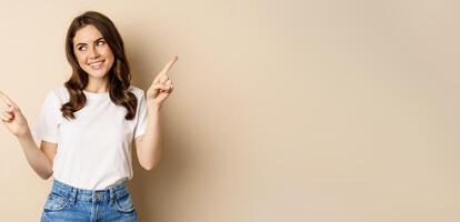 Happy authentic girl smiling, pointing fingers sideways, showing left and right banner, demonstrating promo, standing against beige background photo