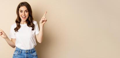 Happy authentic girl smiling, pointing fingers sideways, showing left and right banner, demonstrating promo, standing against beige background photo