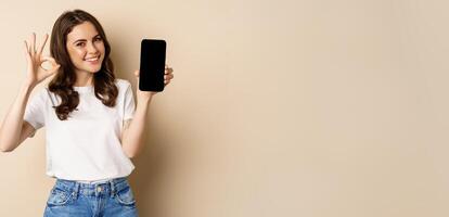 Smiling happy girl showing mobile phone screen, app on smartphone and okay, recommending online store, shopping application, beige background photo