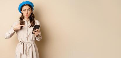 Excited girl with smartphone pointing finger at mobile screen, looking amazed, wow intrigued face expression, beige background photo