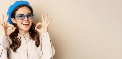 Image of happy smiling woman in stylish sunglasses, showing okay signs in approval, recommending eyewear store, beige background photo
