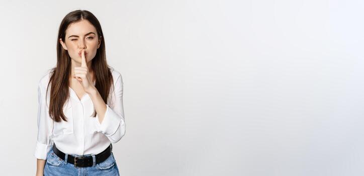 sonriente hermosa mujer callar, haciendo Cállate shh firmar, prensa dedo a labios y parpadeo, ocultación secreto, en pie en blusa y pantalones terminado blanco antecedentes foto