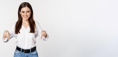Confident smiling woman pointing fingers down, showing logo, banner below, recommending click on link, standing over white background photo