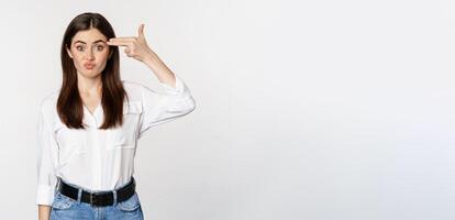 Image of annoyed young woman showing finger gun gesture over head, bothered by smth, tired, standing over white background photo