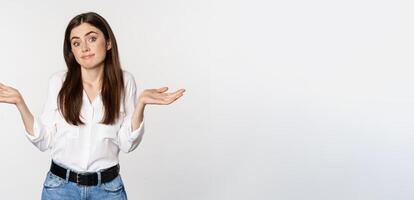 I dont know. Clueless young woman shrugging shoulders and looking unaware, standing over white background photo