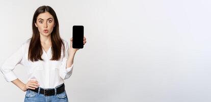 Enthusiastic corporate woman demonstrating website, mobile phone screen, showing application and saying wow, standing over white background photo
