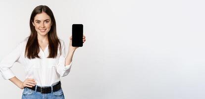Confident woman in corporate clothes, showing smartphone screen, mobile interface of an application, standing over white background photo