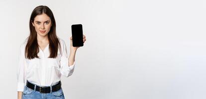 Confident woman in corporate clothes, showing smartphone screen, mobile interface of an application, standing over white background photo