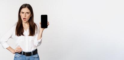 Shocked and frustrated woman showing mobile phone screen app, smartphone application, looking insulted, standing over white background photo