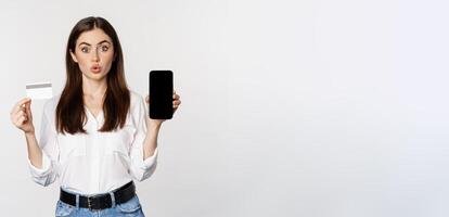 Happy woman showing credit card and smartphone screen, concept of online shopping, buying in app, standing over white background photo