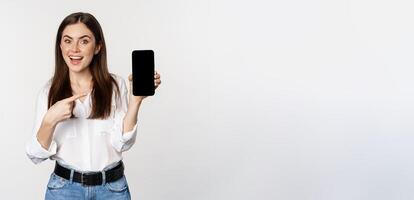 Happy beautiful woman smiling, pointing at mobile phone screen, showing advertisement, website, standing over white background photo