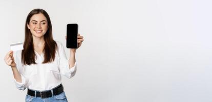 Portrait of female model showing credit card with smartphone screen, recommending application, standing over white background photo