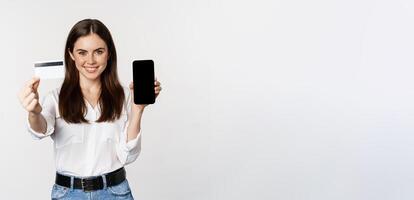 Portrait of female model showing credit card with smartphone screen, recommending application, standing over white background photo