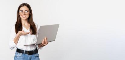 Portrait of woman in glasses holding laptop, pointing at screen, showing her work on computer, standing over white background photo
