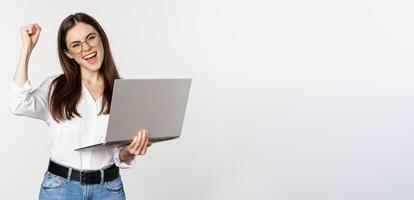 Enthusiastic office woman, businesswoman holding laptop and shouting with joy, celebrating and rejoicing, standing over white background photo