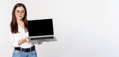 Young woman in glasses showing laptop screen, demonstrating promo on computer, website or store, standing over white background photo