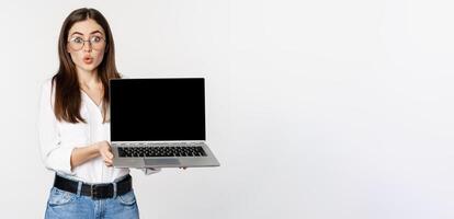 Portrait of cute girl in glasses, student showing laptop screen with amazed face, standing over white background photo