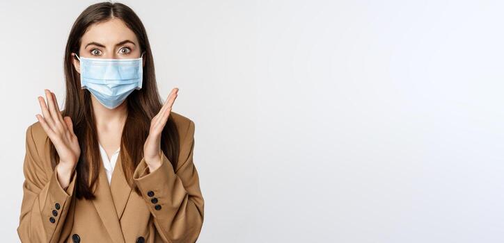 lugar de trabajo y pandemia concepto. conmocionado negocio mujer en cara médico mascarilla, jadeando, mirando sorprendido y preocupado a cámara, en pie terminado blanco antecedentes foto