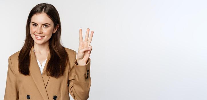 retrato de corporativo mujer, vendedora demostración número Tres dedos y sonriente, en pie en traje terminado blanco antecedentes foto