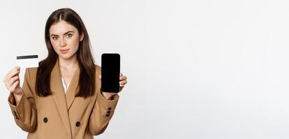 Businesswoman showing mobile phone screen and credit card, demonstrating application on smartphone, standing in suit over white background photo