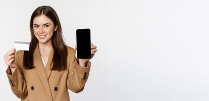 Businesswoman showing mobile phone screen and credit card, demonstrating application on smartphone, standing in suit over white background photo